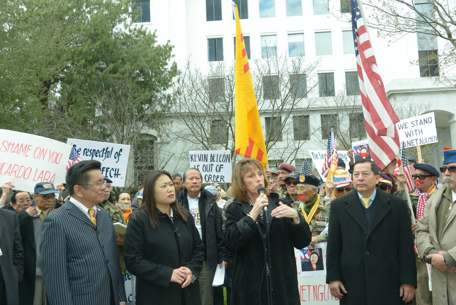 Phat Bui stands in the front row beside a woman speaking into a microphone at an outdoor rally; the crowd behind holds flags and signs including “We stand with Janet Nguyen”