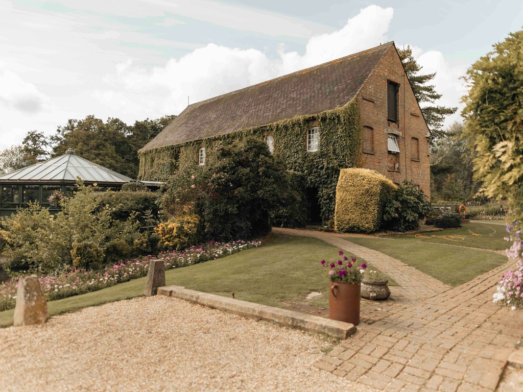 Ivy-covered brick barn wedding venue with manicured gardens, a brick pathway, and a glass conservatory on a sunny day