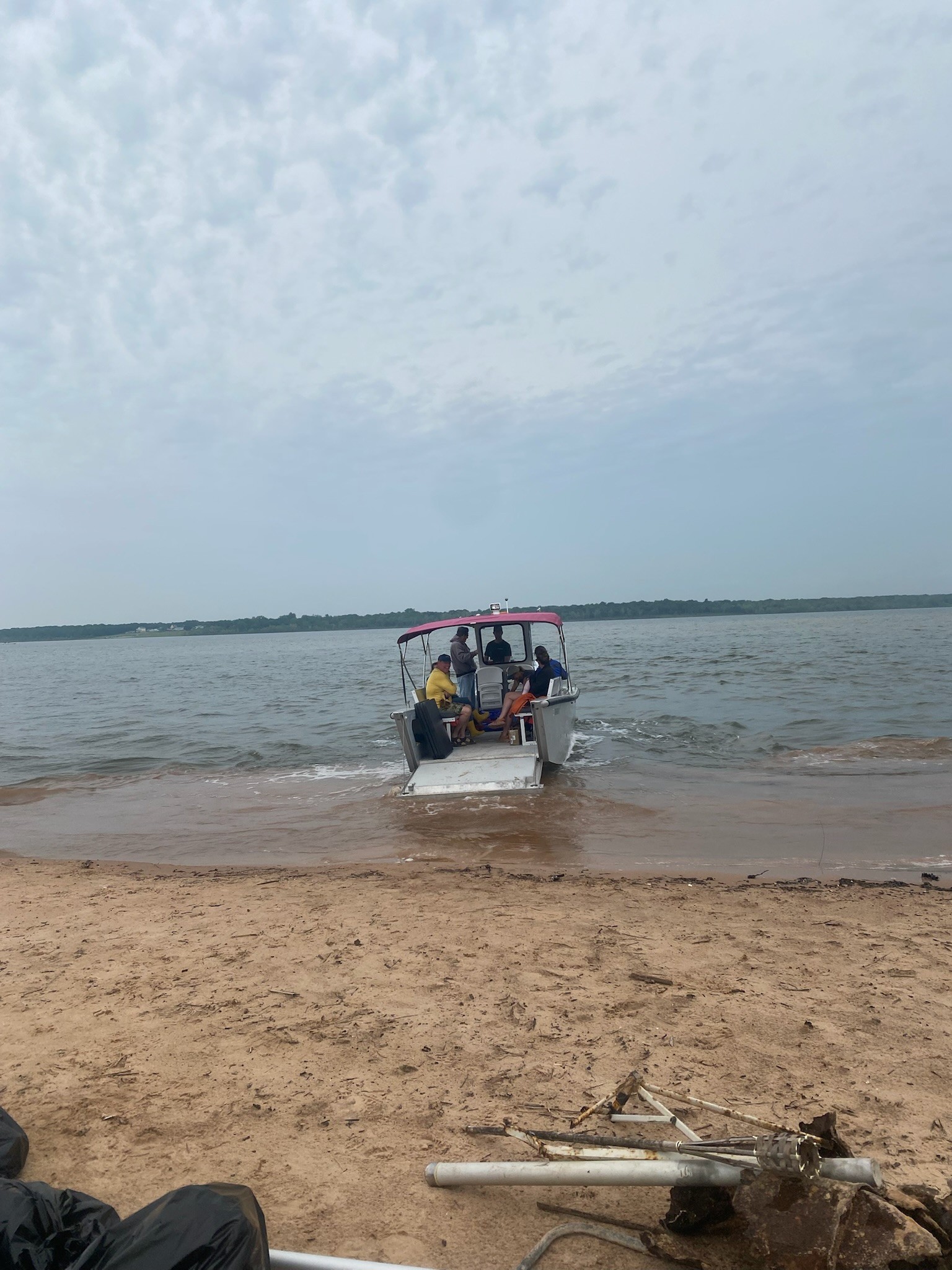 A small motorboat with a red canopy approaches a sandy shore under a cloudy sky, with passengers onboard and the calm waters of the lake in the background.