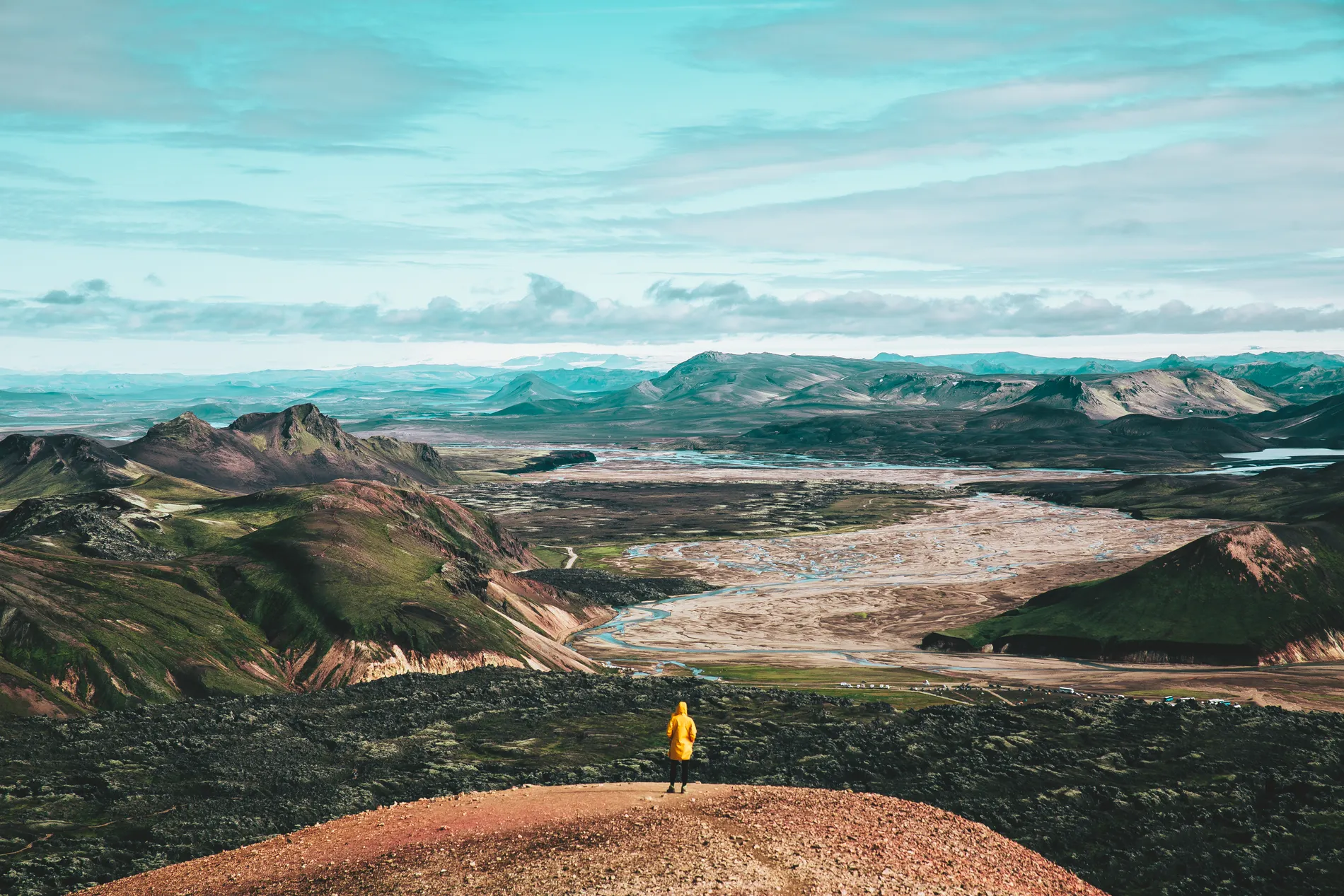 A figure in a yellow jacket on a red crater rim above a wide volcanic plain