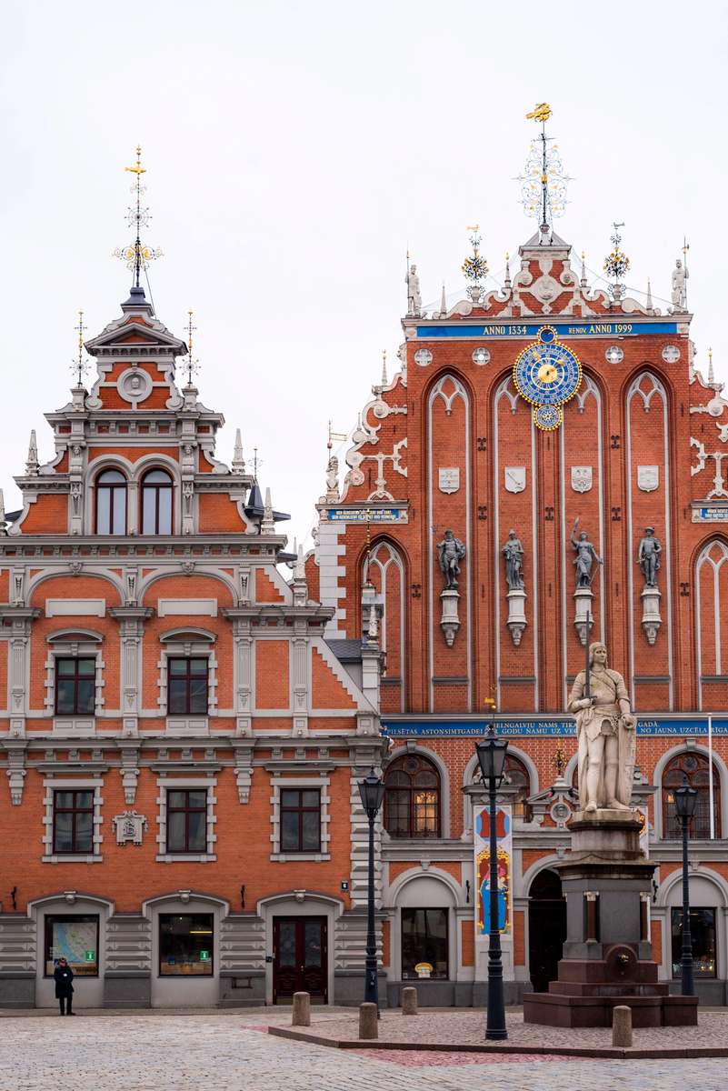 Ornate red and white buildings with golden domes and a statue on a pedestal in a paved public square under an overcast sky.