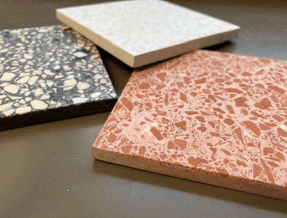 Trois carreaux carrés en terrazzo recyclé posés sur une surface sombre : un rose-corail, un noir et un blanc, tous incrustés d'éclats multicolores de céramique broyée
