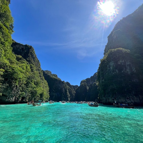 Turquoise water bay surrounded by steep, lush cliffs under a clear sunny sky; numerous boats scattered across the water.