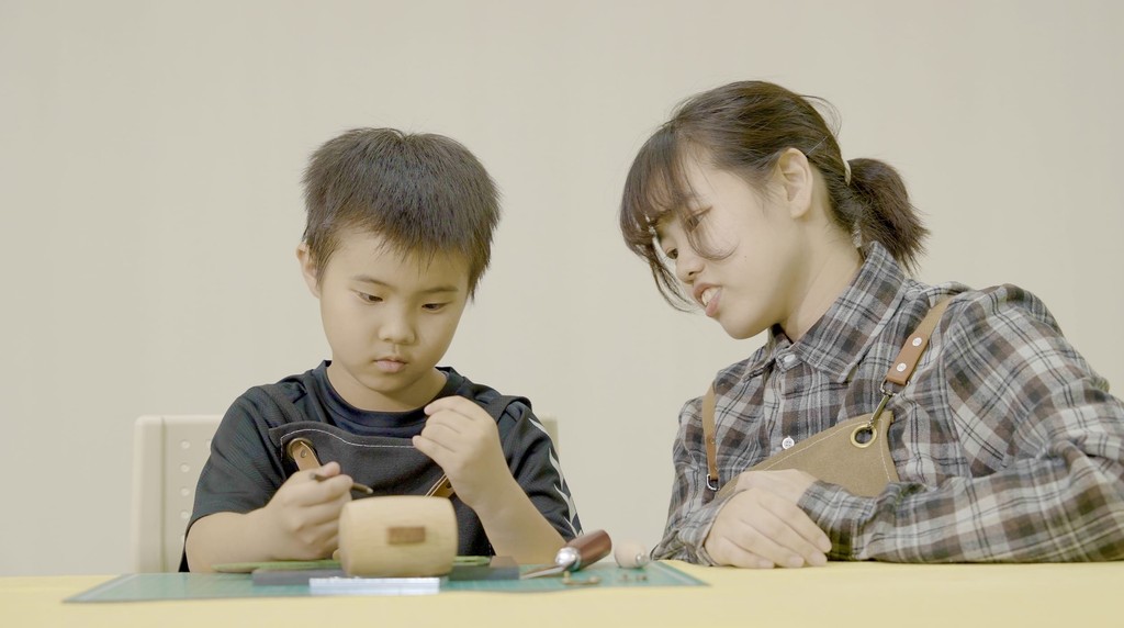 An instructor at our kids academy provides focused, one-on-one guidance to a young student during a hands-on craft workshop. The boy concentrates intently on his project at a yellow table while the teacher leans in to assist.