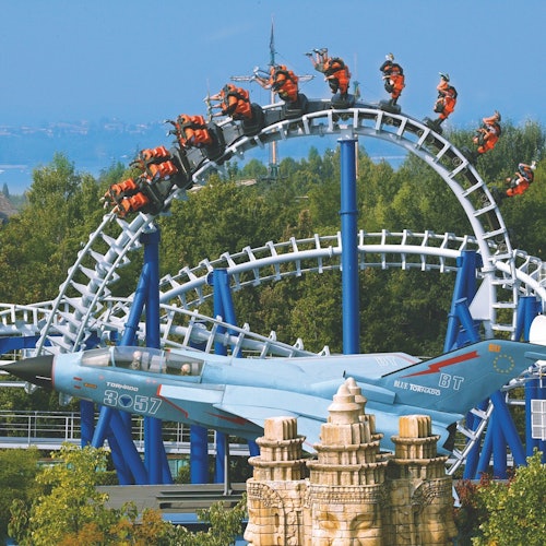 אנשים רוכבים על רכבת הרים שעושה לולאה מעל מטוס סילון לתצוגה, מוקף בעצים עם נוף עירוני מרוחק ברקע.