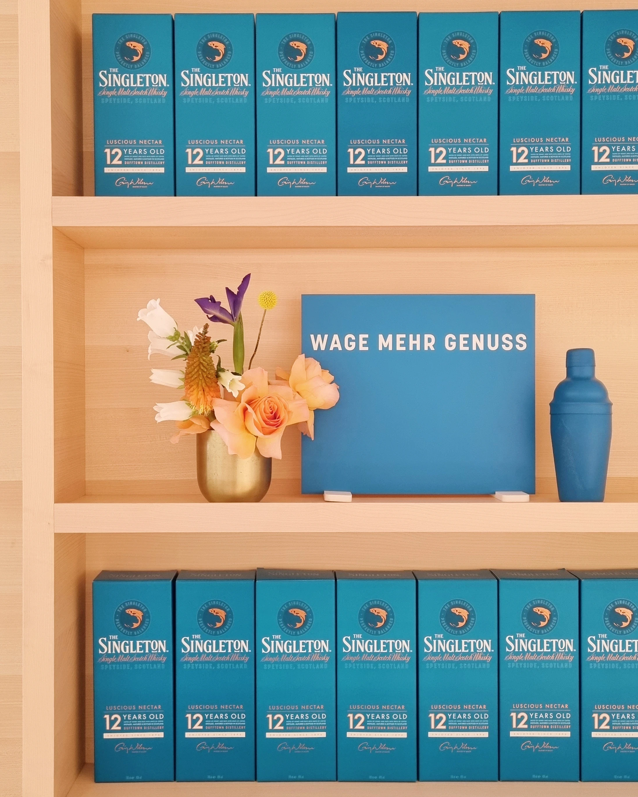 Wooden shelf with rows of teal Singleton boxes around a Wage mehr Genuss sign and flowers.