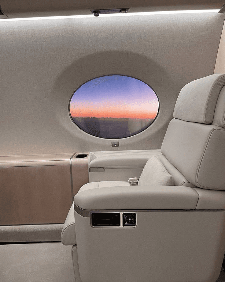 Interior of private jet with luxurious leather seat, porthole window showcasing scenic sunset.