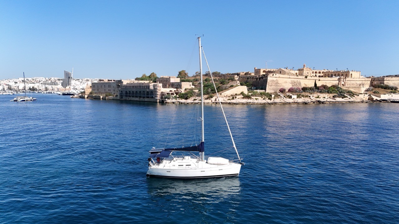 Aerial drone shot of a white Beneteau Oceanis Clipper 373 sailboat with sails furled, floating on calm deep-blue water in Grand Harbour, Valletta, Malta. Historic limestone fortifications and waterfront buildings are visible in the background under a clear blue sky.