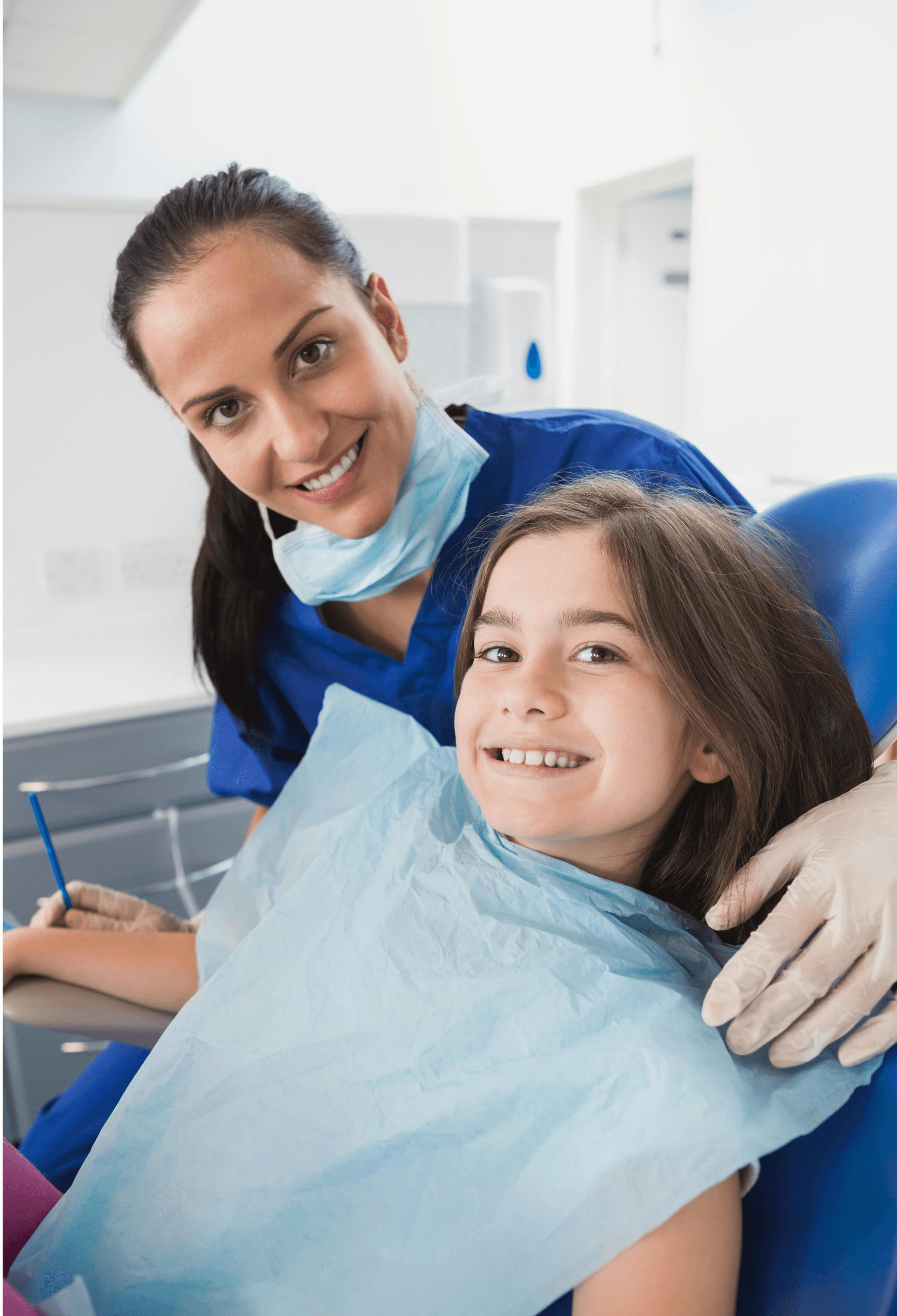 A dentist in a blue coat and mask smiling alongside a young girl in a dental chair, both looking happy and comfortable during the dental visit.