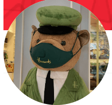 The image shows a teddy bear in a green uniform and "Harrods" face mask, set in a retail environment.