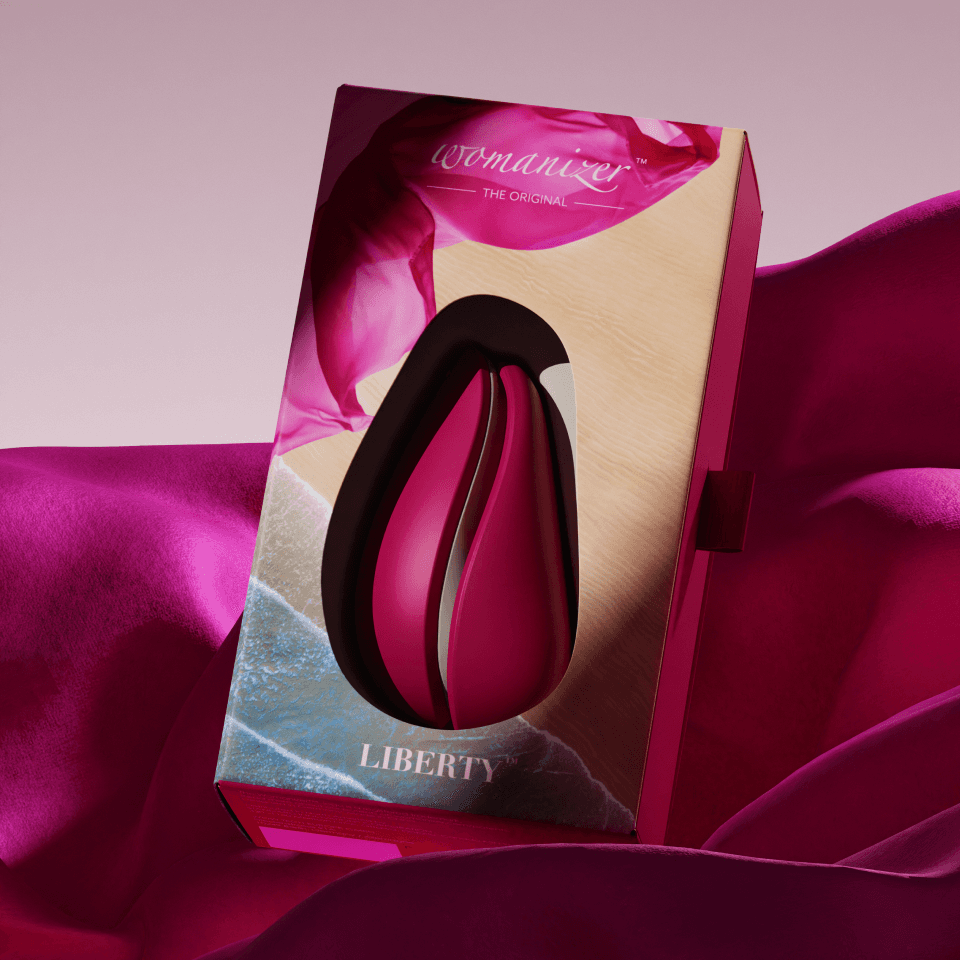 Womanizer „Liberty" Detailansicht – Verpackung in Beerentönen mit Insel- und Küsten-Artwork, eingebettet in fliessende Magenta-Seide.