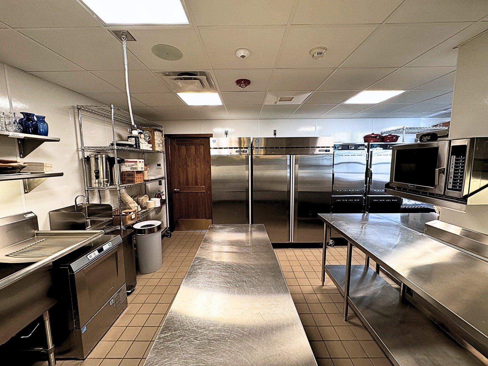 Commercial kitchen at Grace Trinity Church featuring stainless steel appliances, large refrigerators, industrial microwave, and shelving with kitchen supplies.