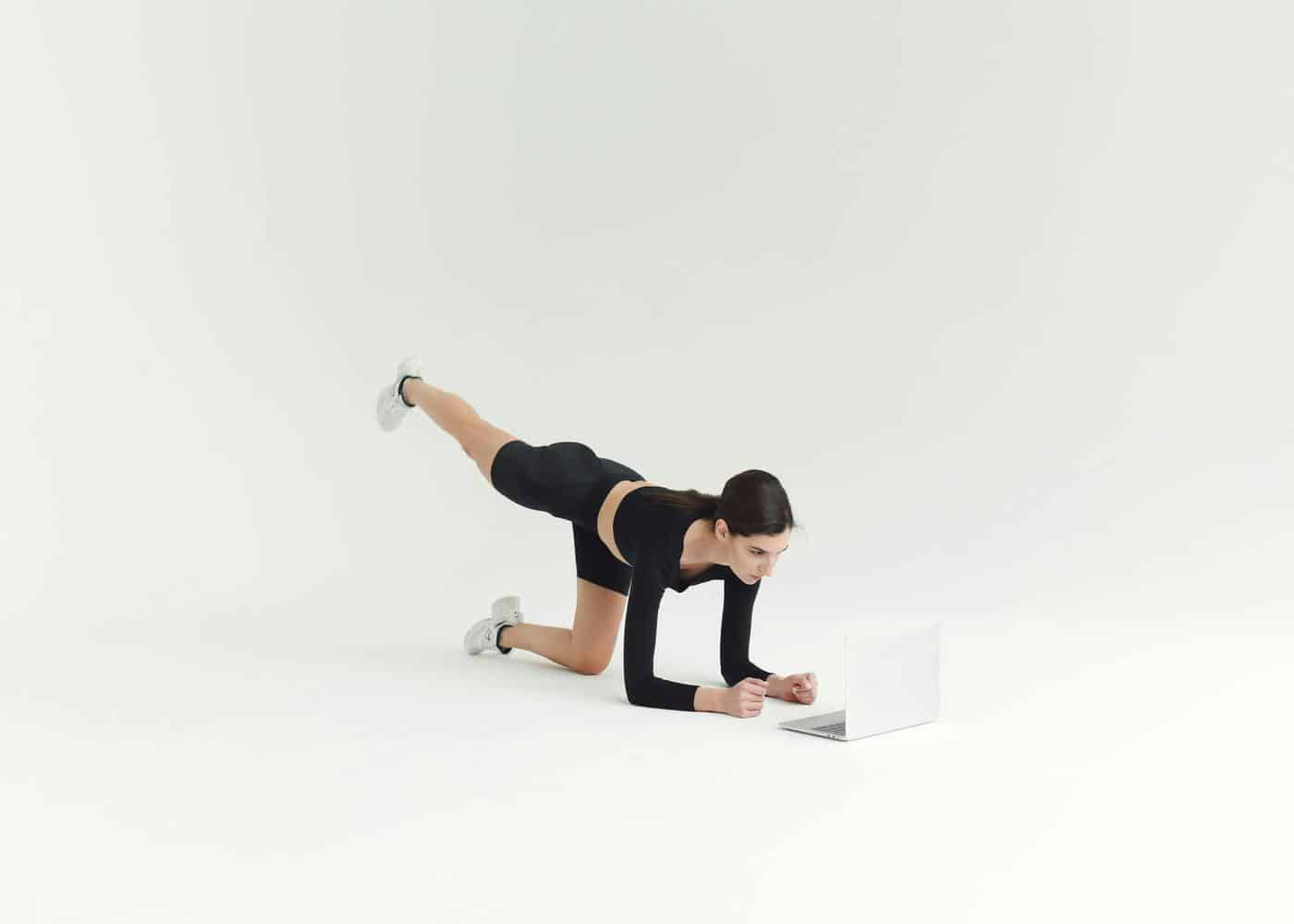 Woman working out doing a plank on her forearms while raising her leg and looking at a laptop in front of her