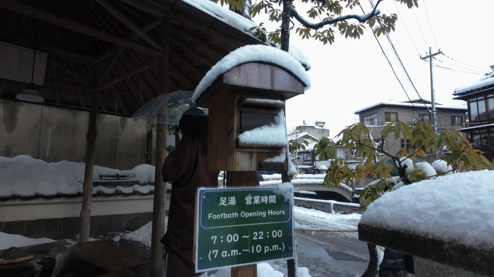 Bilingual sign at the Kinosaki station footbath reading Footbath Opening Hours 7:00 to 22:00, snow on the roof.