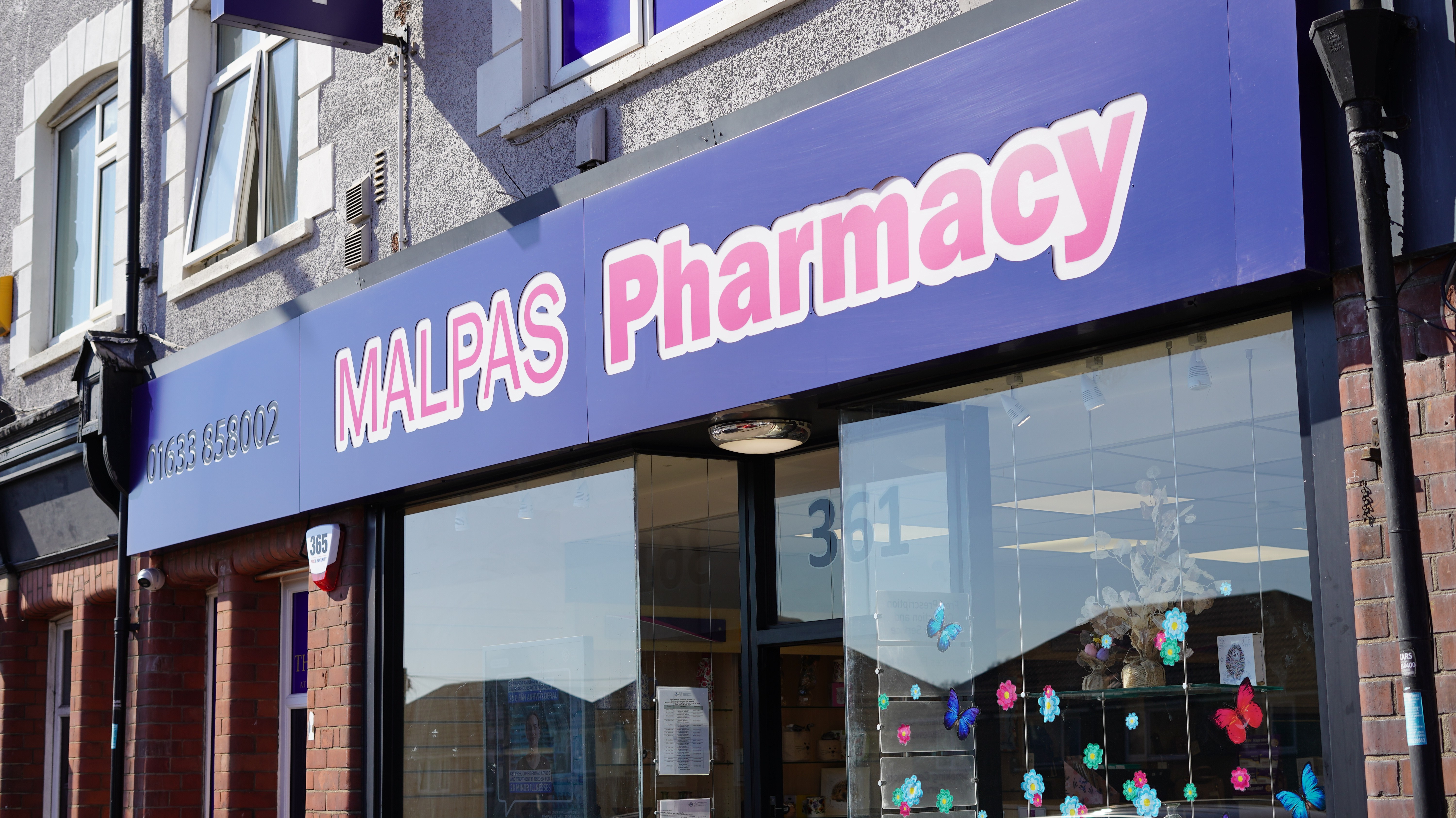 Exterior of Malpas Pharmacy in Newport, South Wales, showing a blue shopfront with large pink-and-white “Malpas Pharmacy” signage above the entrance and front windows. The glass displays colourful butterfly and flower decorations, with the number 361 visible on the door.