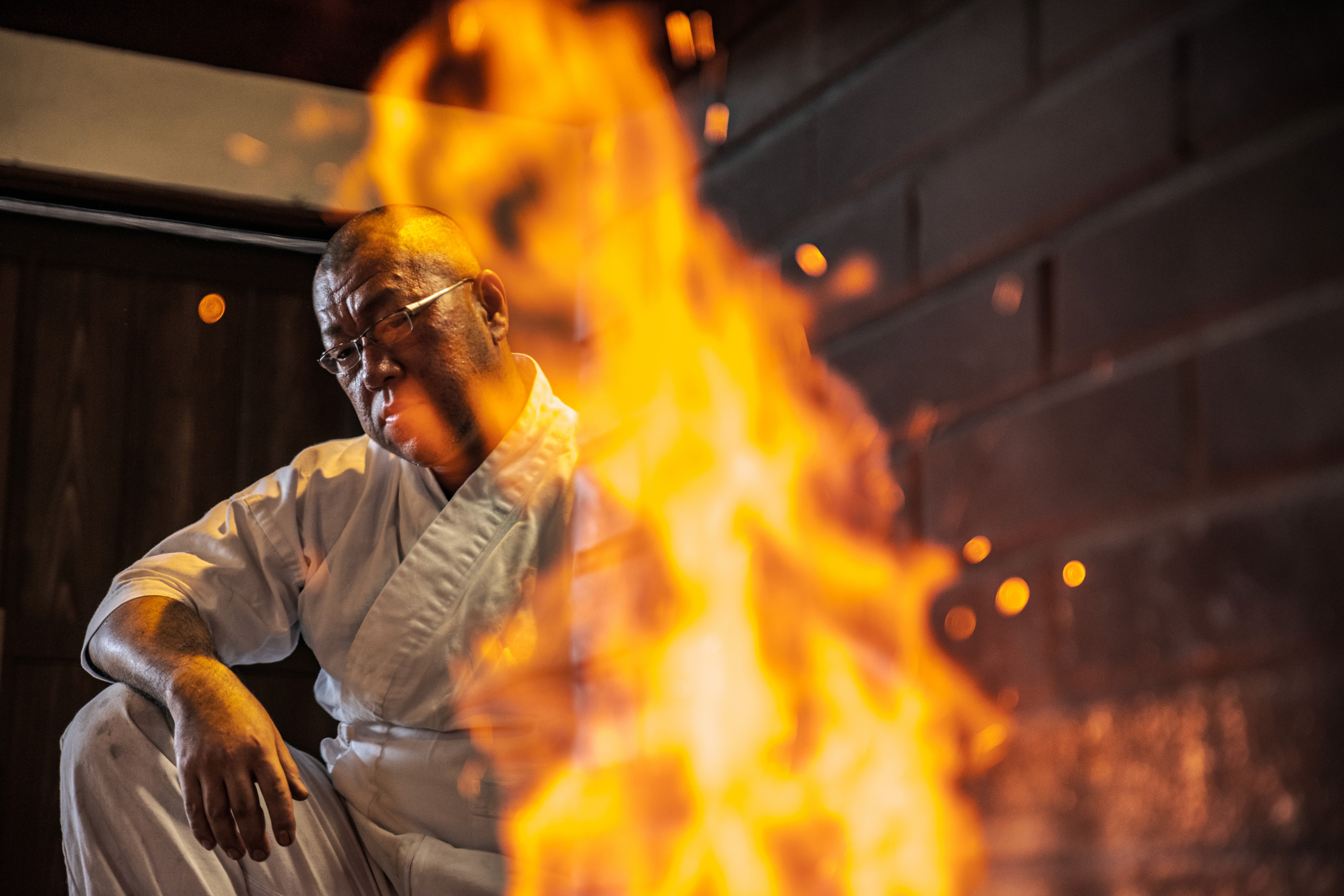 A swordsmith stands behind a towering flame in his workshop