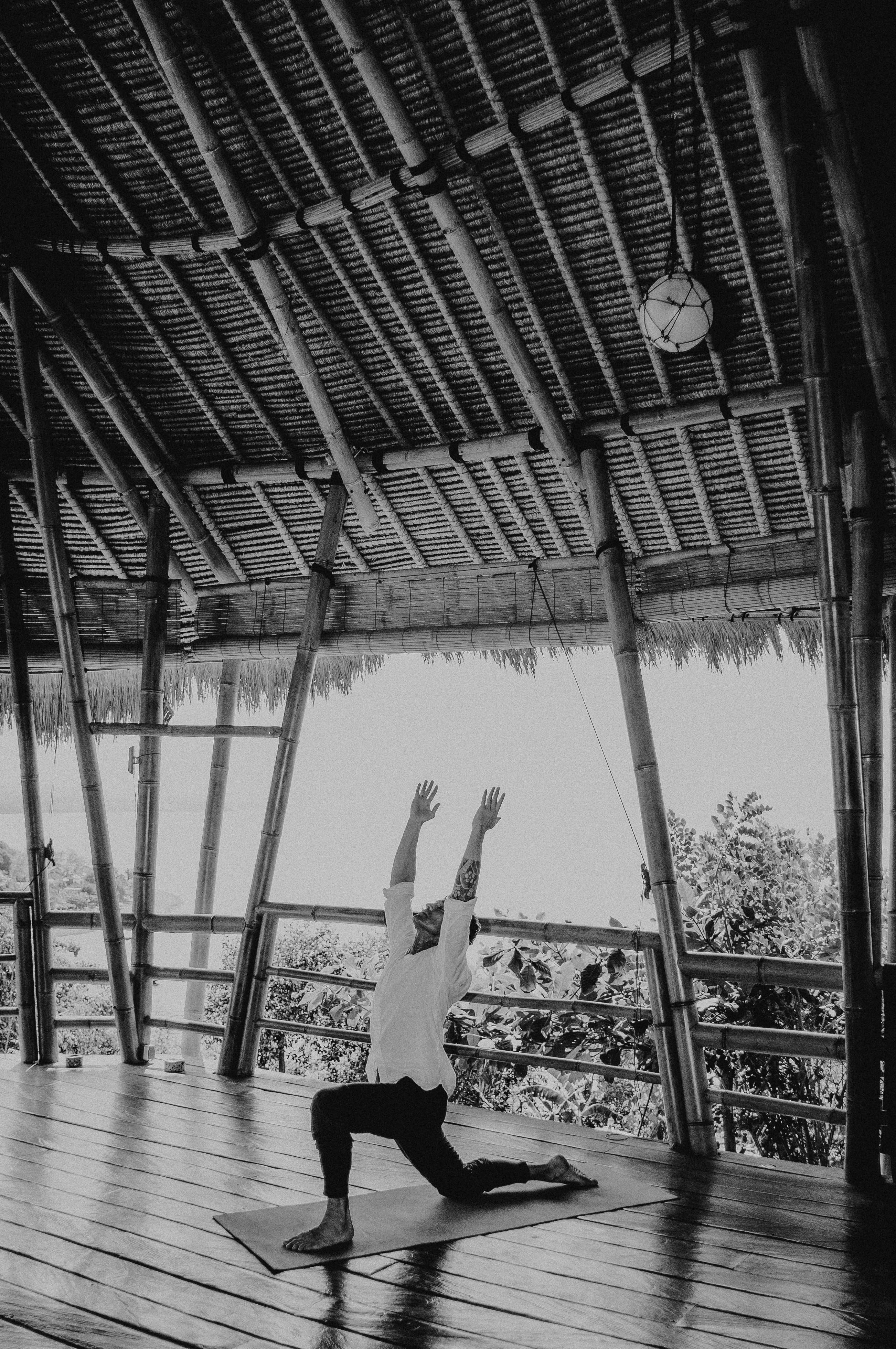 A person practices yoga on a wooden deck with a high bamboo ceiling, surrounded by lush greenery under natural light.