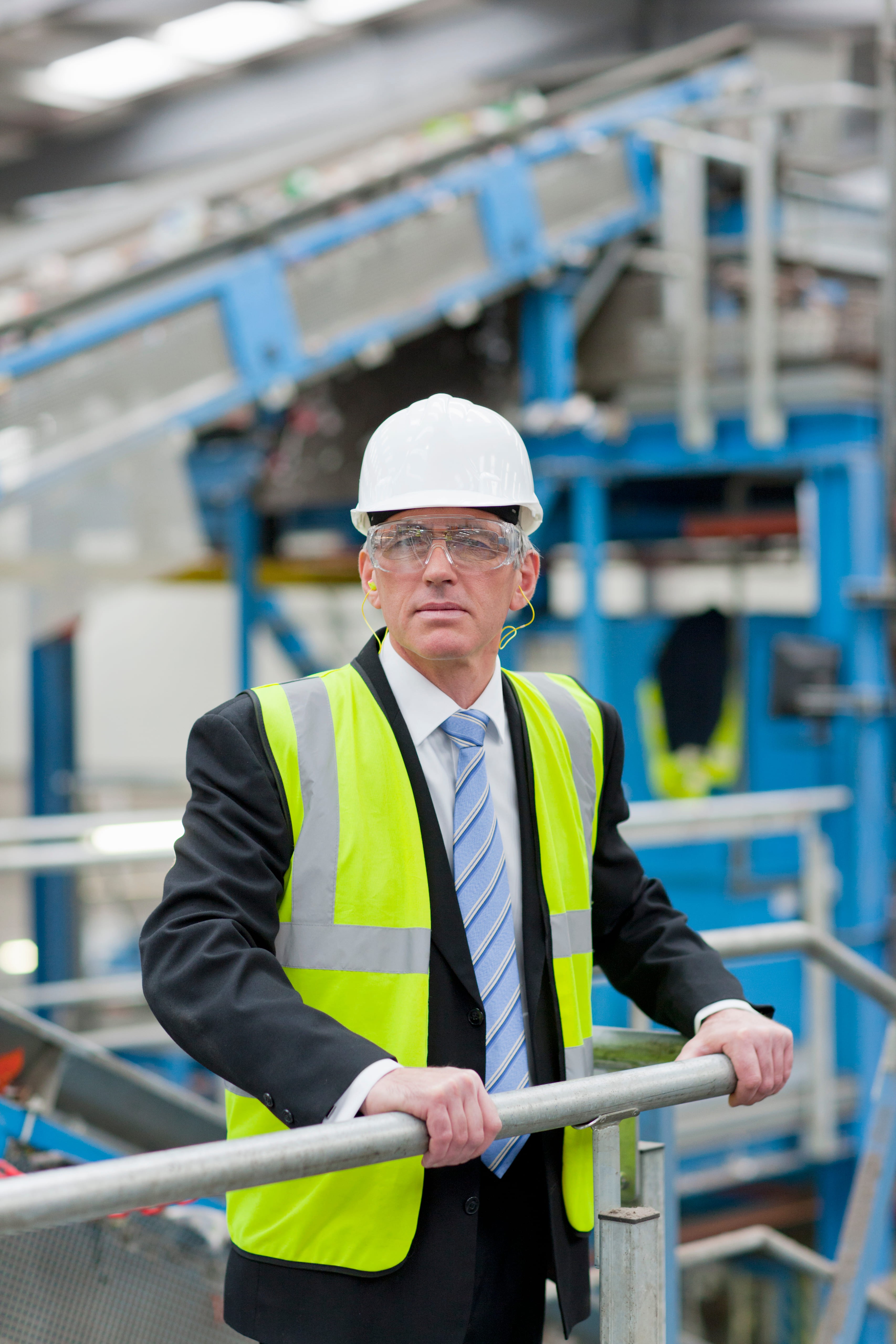 Profissional vestindo capacete e colete fluorescente apoiado em corrimão ambiente industrial.