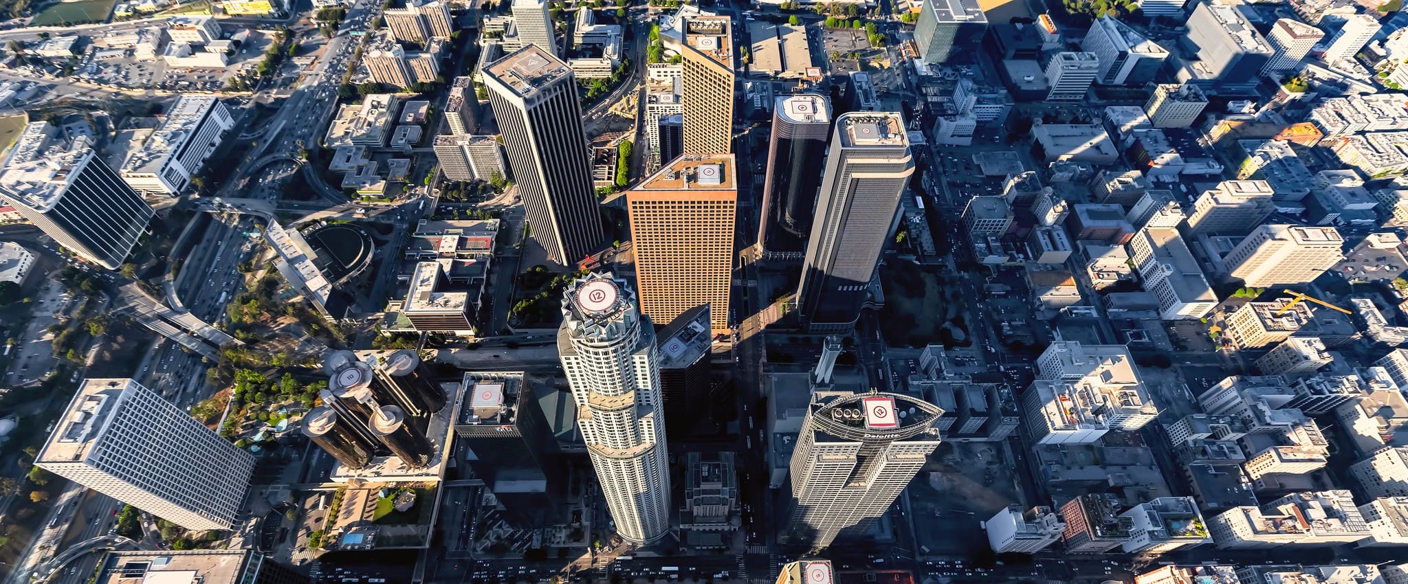 A high-angle aerial view looking straight down at the downtown Los Angeles skyline, showcasing dense skyscrapers with helipads on their roofs and surrounding city streets.