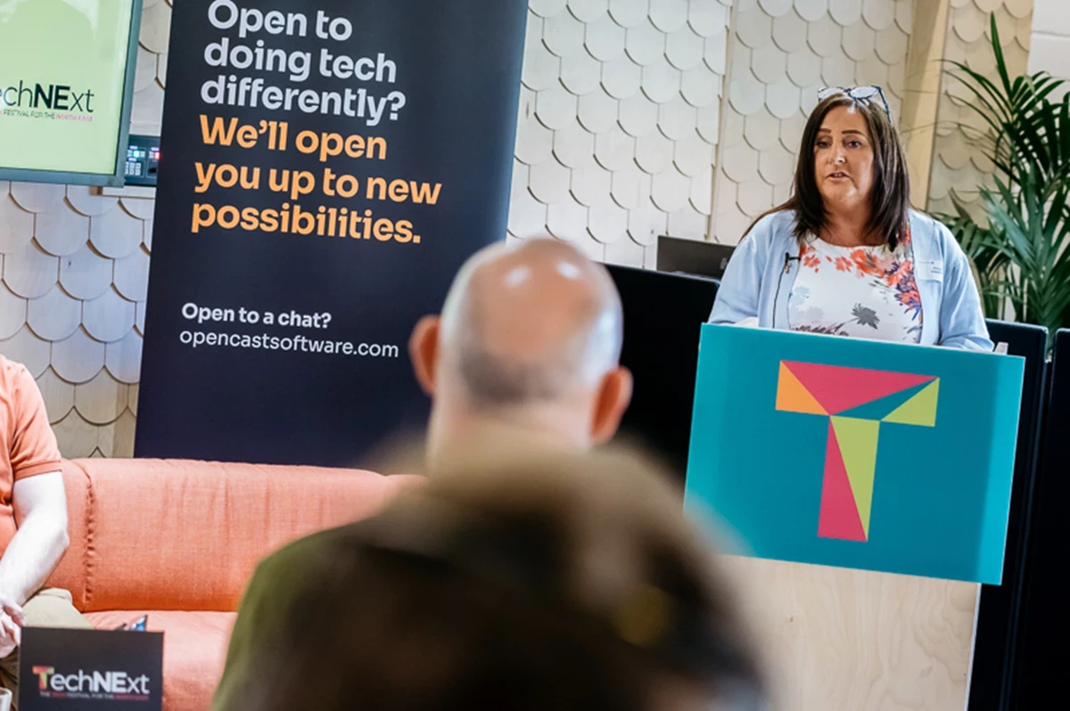 Person standing behind a podium with a colorful geometric logo, speaking to an audience, with a banner in the background that reads ‘Open to doing tech differently? We’ll open you up to new possibilities.’