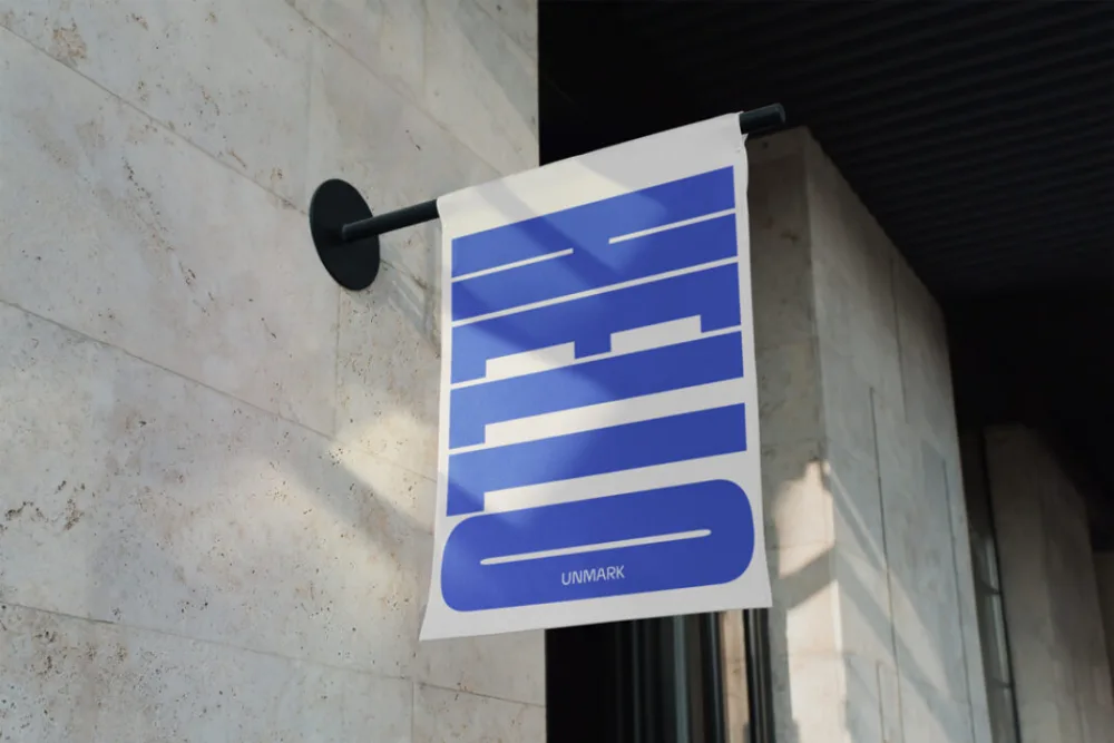 Wall-mounted vertical banner mockup featuring a fabric sign with a modern black bracket. The banner is white with bold blue lettering, displayed against a textured beige stone wall in natural daylight.