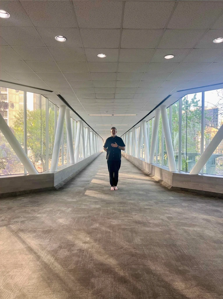 A person stands in a long, bright hallway with glass walls and modern architecture. Soft lighting illuminates the space.