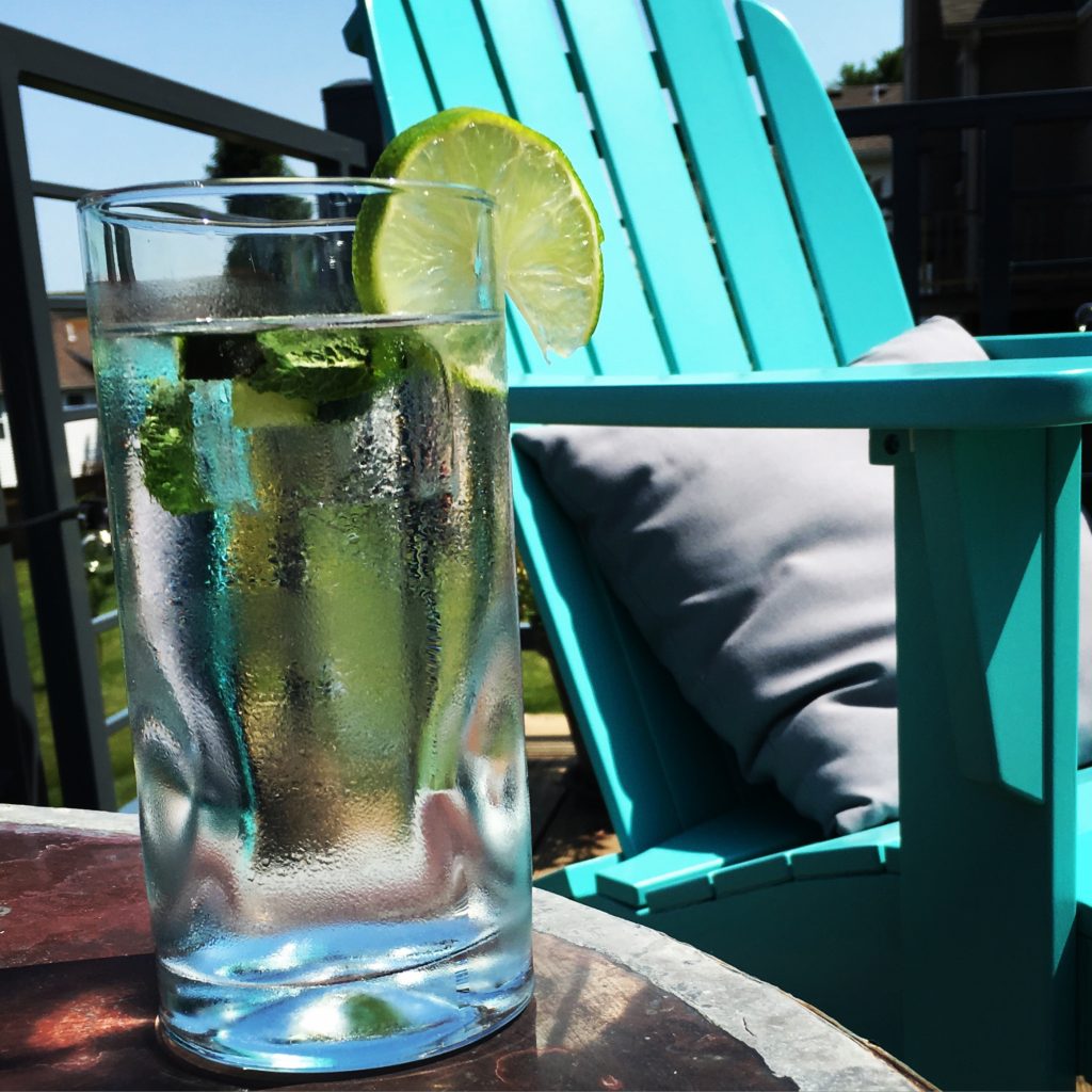 A refreshing glass of sparkling water with lime slices sits on a sunlit patio table, next to a vibrant turquoise Adirondack chair, creating a relaxing outdoor setting.