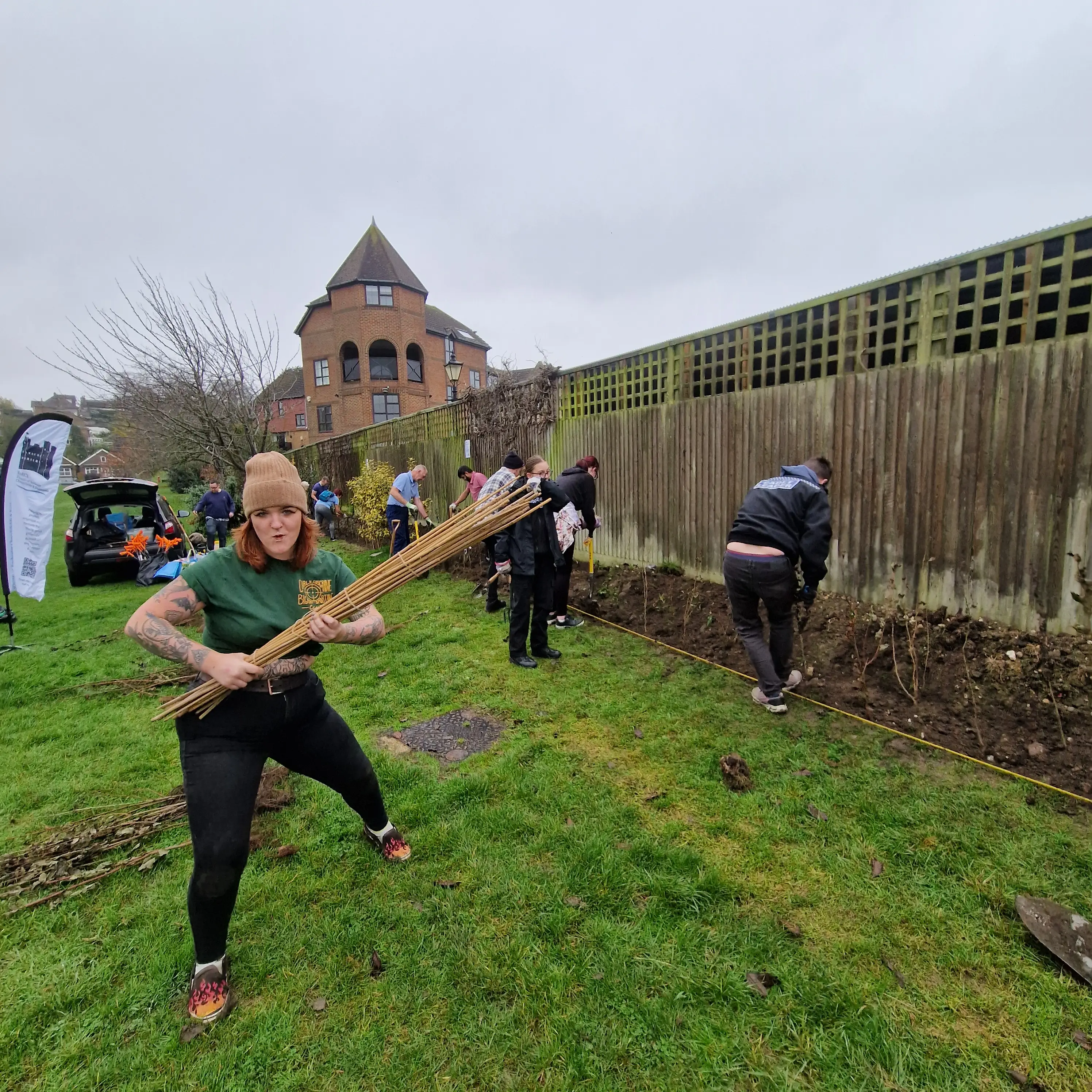 Three people work in a garden, using tools near a fence, with a house visible in the background under cloudy skies.