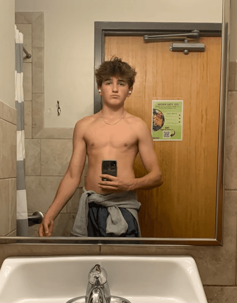 A man with short hair and a beard takes a mirror selfie in a dimly lit bathroom, wearing a green t-shirt and holding a smartphone.