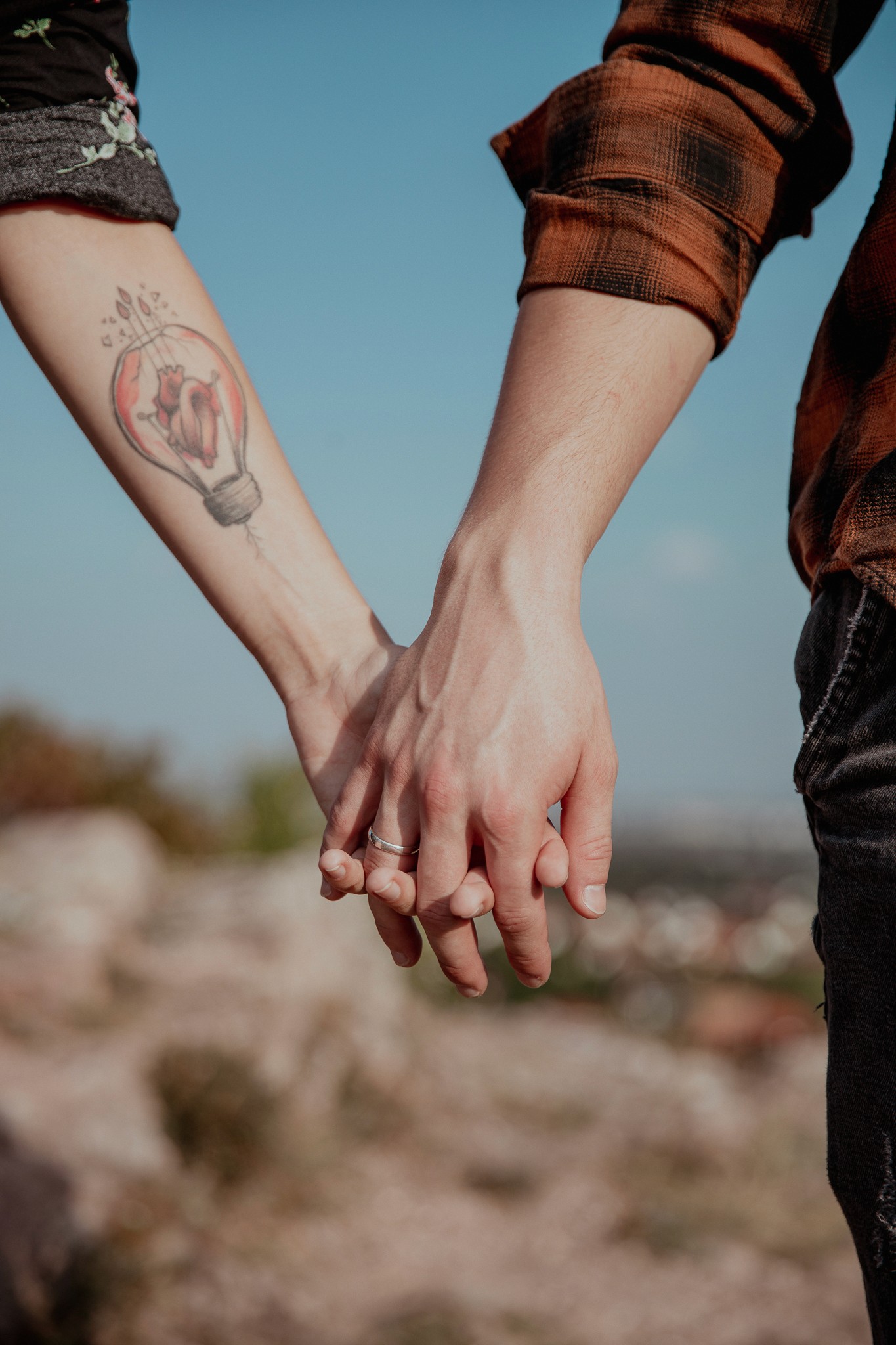 Két ember kézfogását mutató fotó, az egyik karon szívet ábrázoló tetoválással, szabadtéri, napfényes háttérrel.