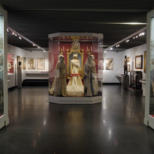 博物馆展览中，历史服饰陈列于玻璃展柜内，背景可见艺术作品和雕塑。