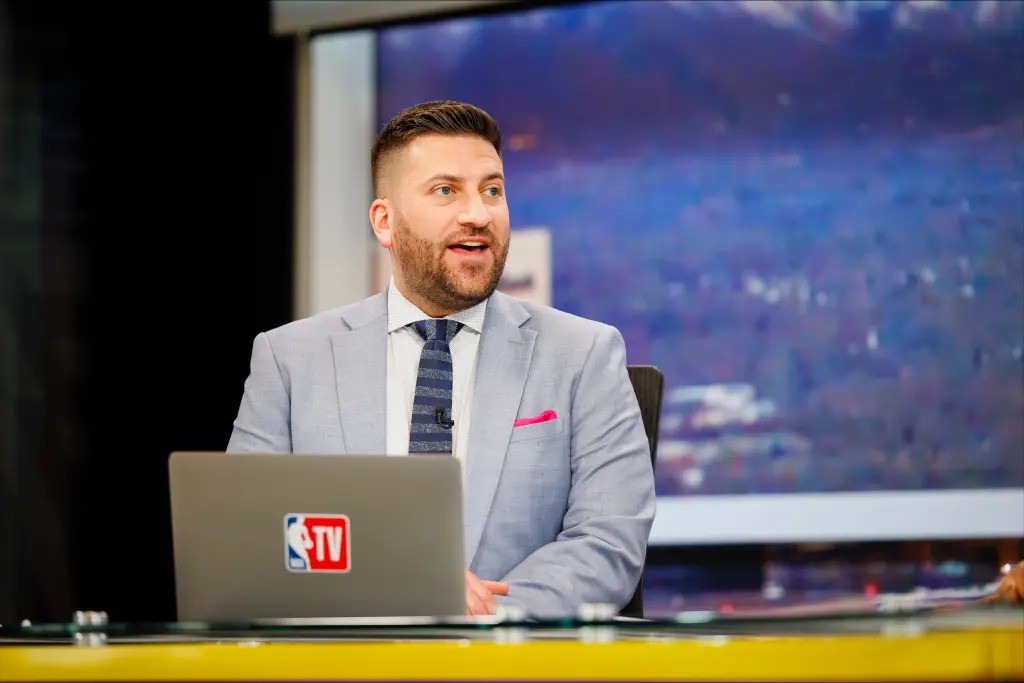 Jared Greenberg in a gray suit with a pink pocket square sits at a desk with an NBA TV laptop, speaking animatedly on a television set with a blurred screen in the background.