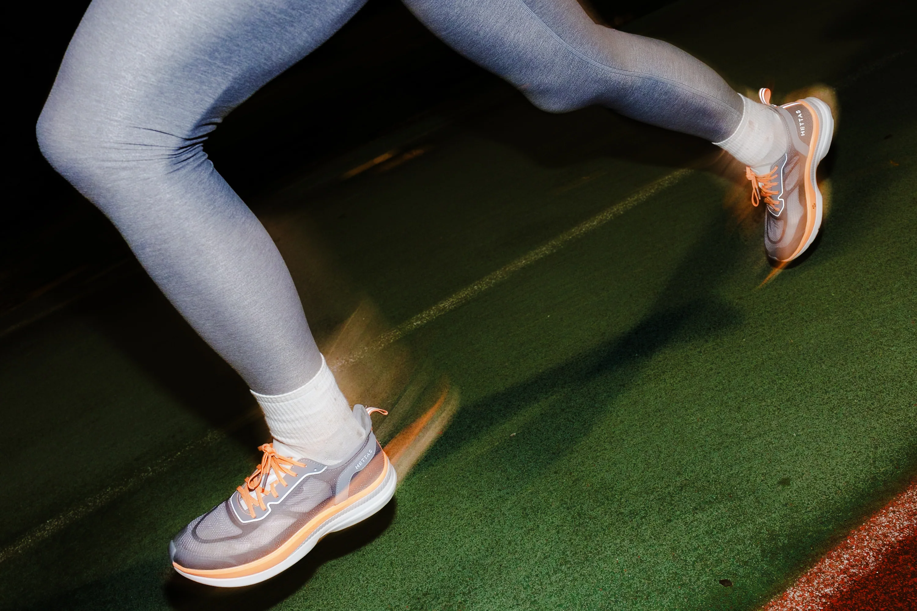 Blurry motion shot of someone running in a pair of charcoal/orange Hettas shoes on a green track