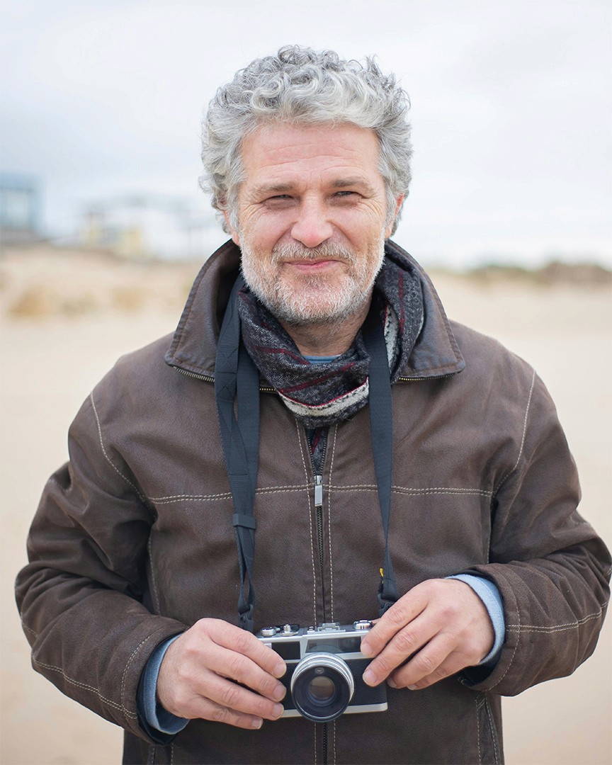 An older gentleman with white hair and a beard in a raincoat standing on a cloudy beach with a camera, smiling