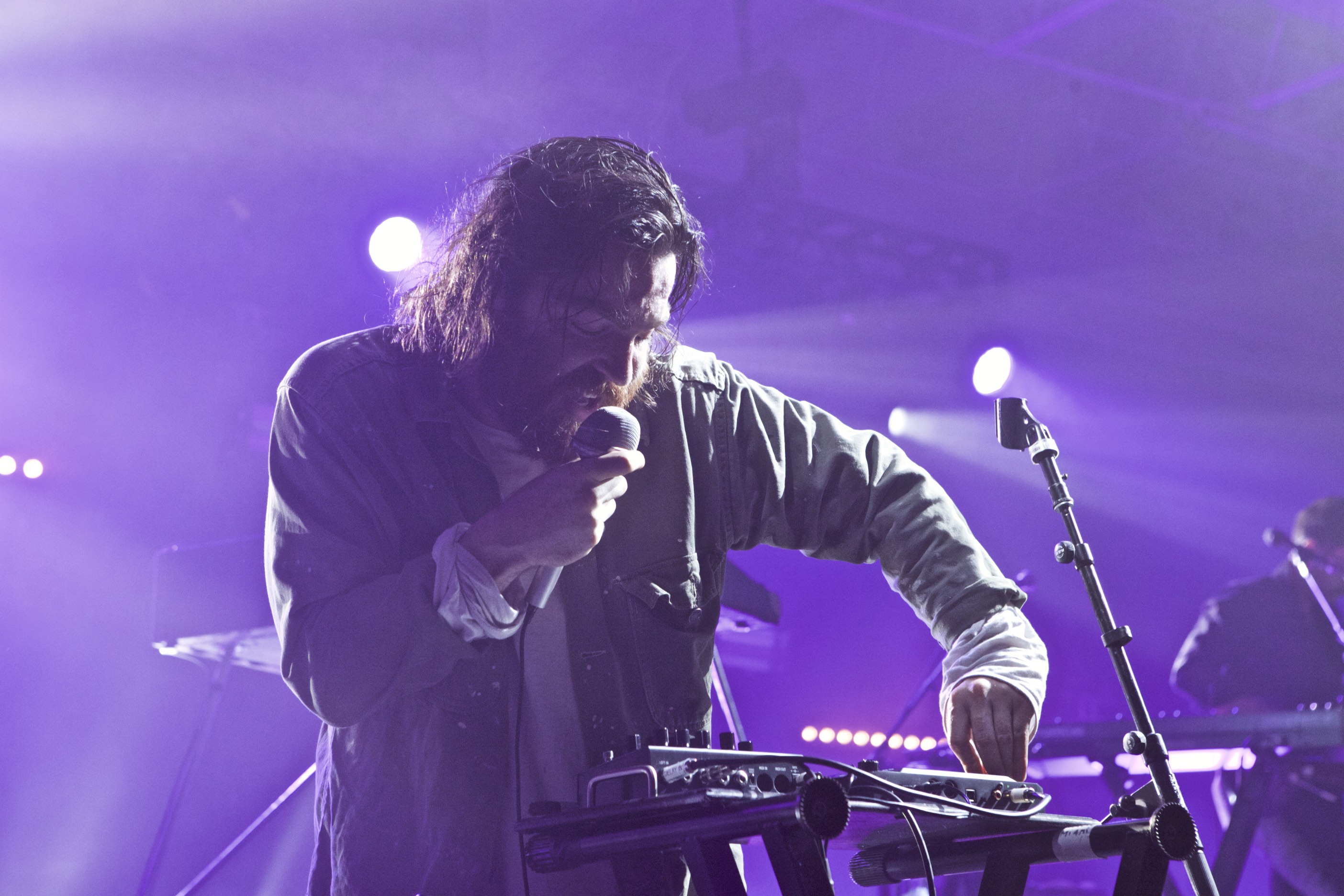 Chet Faker singing into a microphone while playing a small keyboard or synth under purple stage lights.