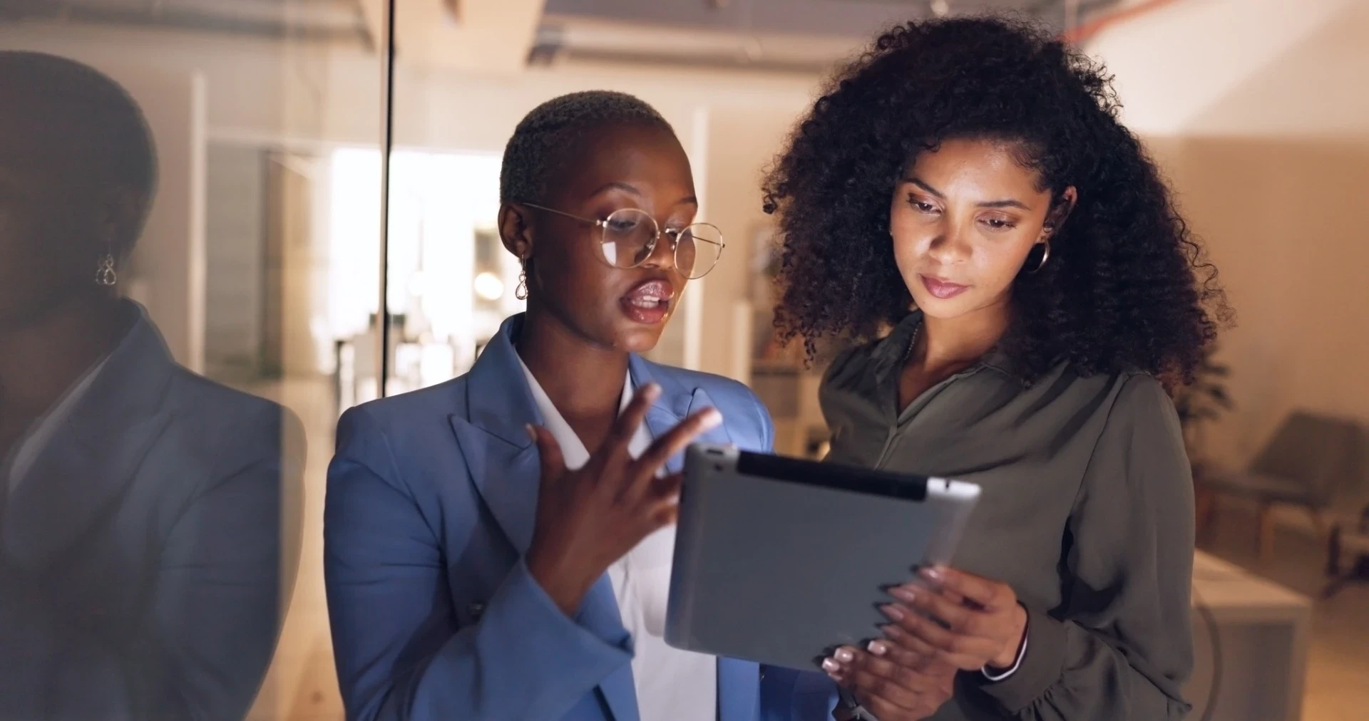 A imagem mostra duas mulheres em um ambiente de trabalho, concentradas enquanto analisam informações em um tablet. Uma delas, à esquerda, usa um blazer azul claro e óculos grandes, e está explicando algo com gestos enquanto fala. A outra, à direita, tem cabelo cacheado e usa uma blusa verde, observando atentamente a tela do dispositivo. O reflexo da luz cria um efeito interessante no vidro ao lado delas. A atmosfera parece ser de colaboração e troca de ideias, com foco na tecnologia e no trabalho em equipe.