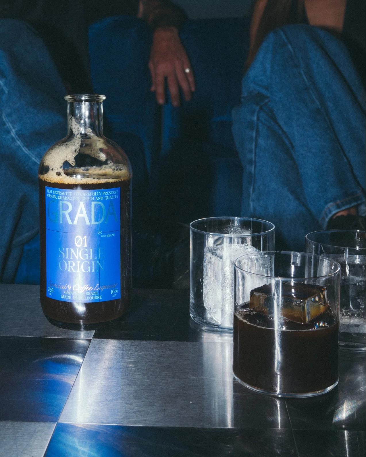 A bottle of dark, single-origin Grada with a blue label sits on a reflective checkered table, surrounded by glasses of iced drinks, with casually dressed individuals relaxing in the background.