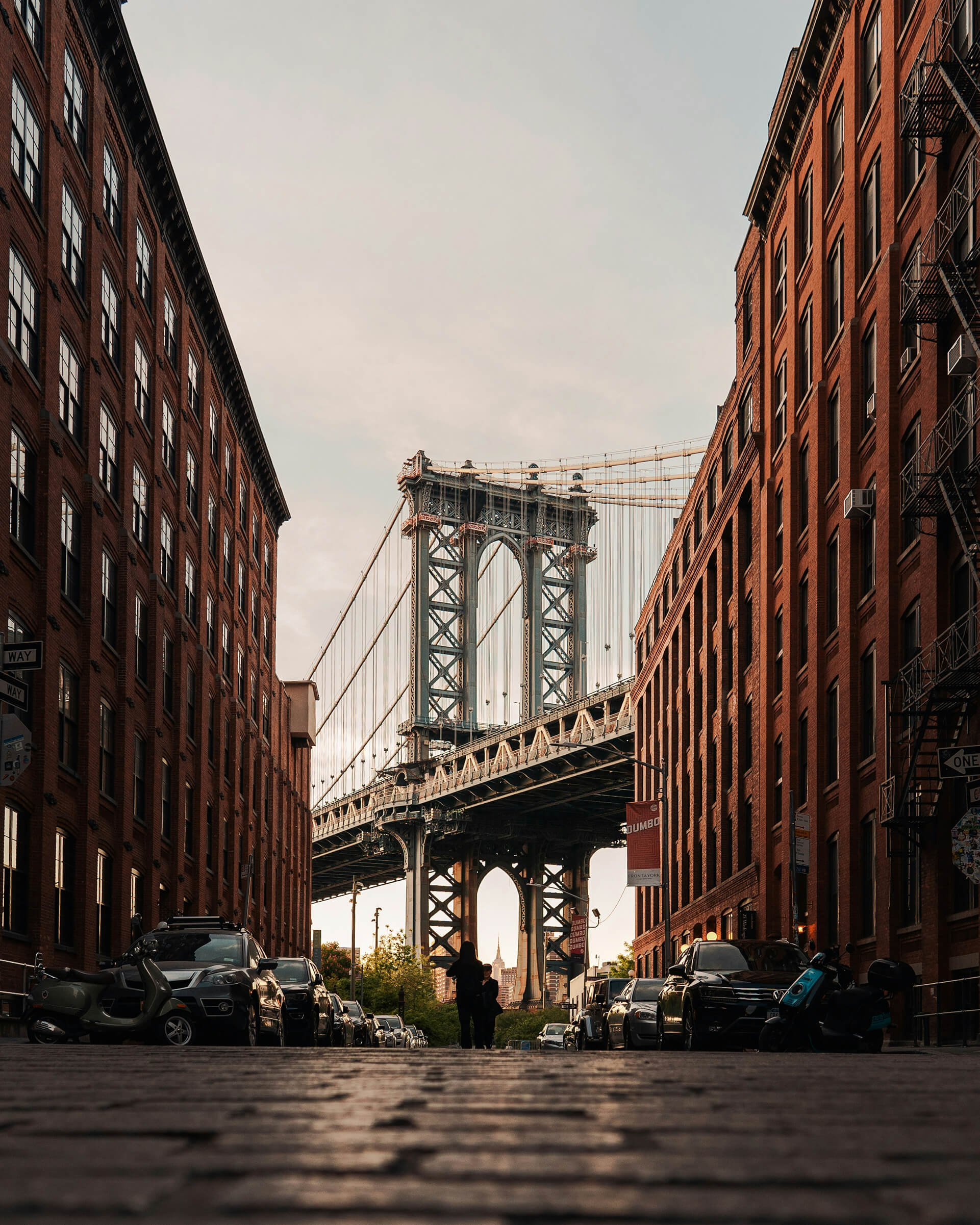 Iconic view of the Manhattan Bridge framed between two red-brick buildings in the DUMBO neighborhood of Brooklyn, New York. A person stands in the cobblestone street at sunset, with the Empire State Building visible through the bridge’s archway in the distance.