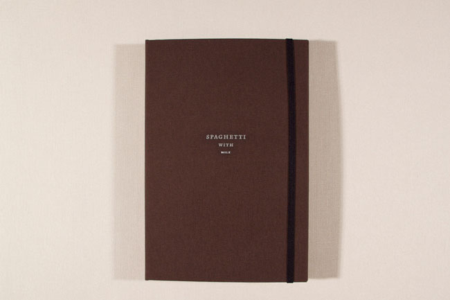 A dark brown notebook with a black elastic band stands upright on a light beige surface. The notebook’s cover features the text "SPAGHETTI WITH MILK" printed in a light gray or silver color.
