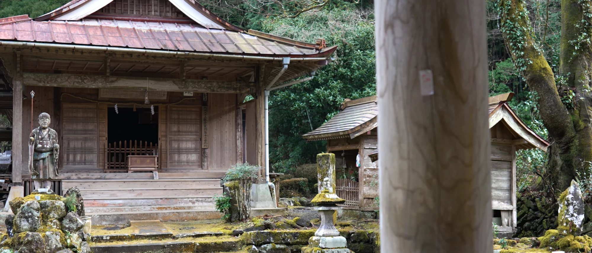 A small timber shrine hall with a weathered stone figure standing before it