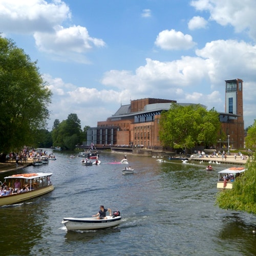 Zahlreiche Boote und Menschen auf einem Fluss neben einem großen Ziegelgebäude, mit Bäumen und einem teilweise bewölkten Himmel im Hintergrund.