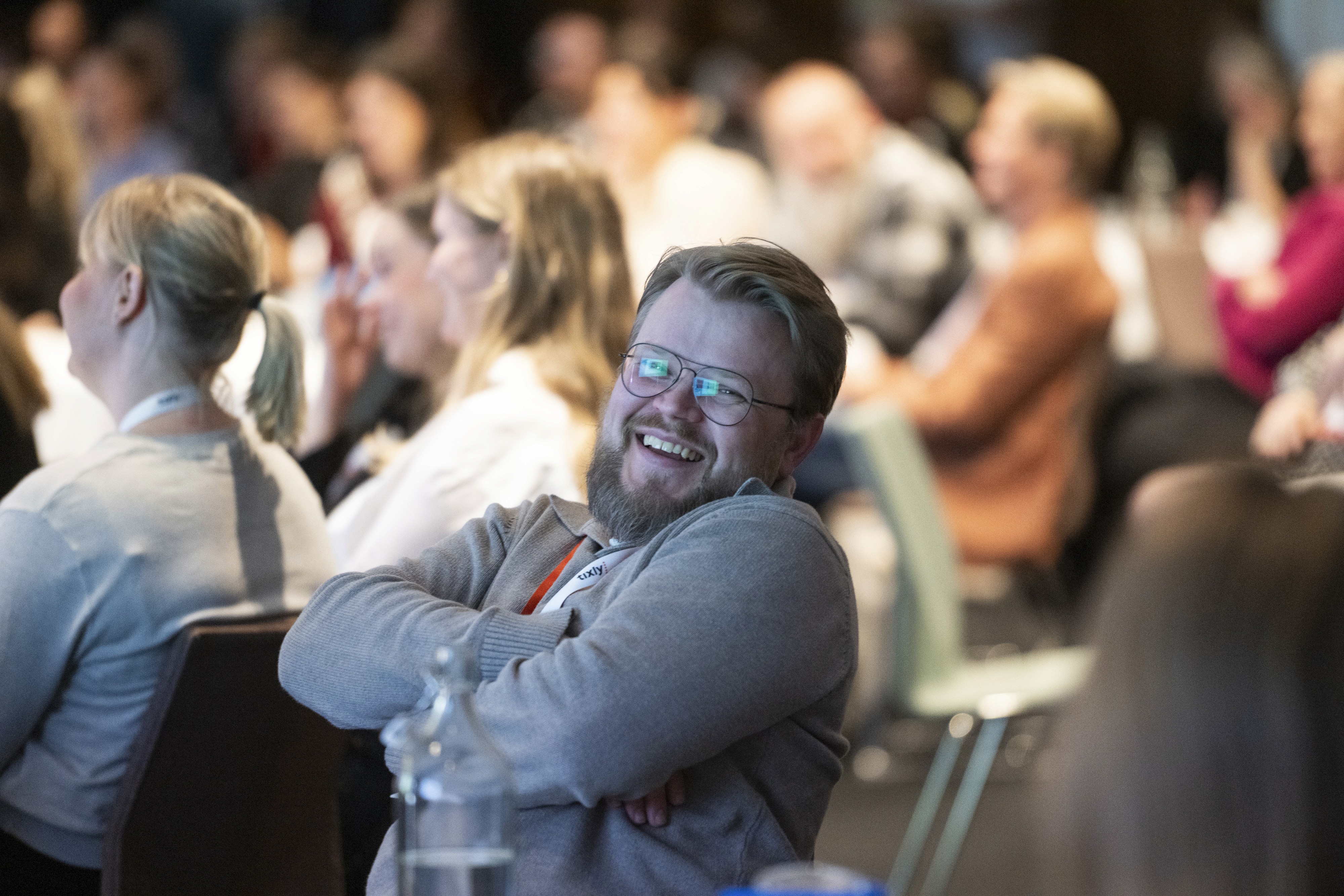 Audience members seated in a conference setting, some engaging while one man looks thoughtfully at the camera.