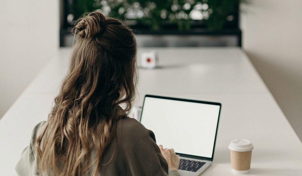 Woman working on laptop at a white desk with a coffee cup.