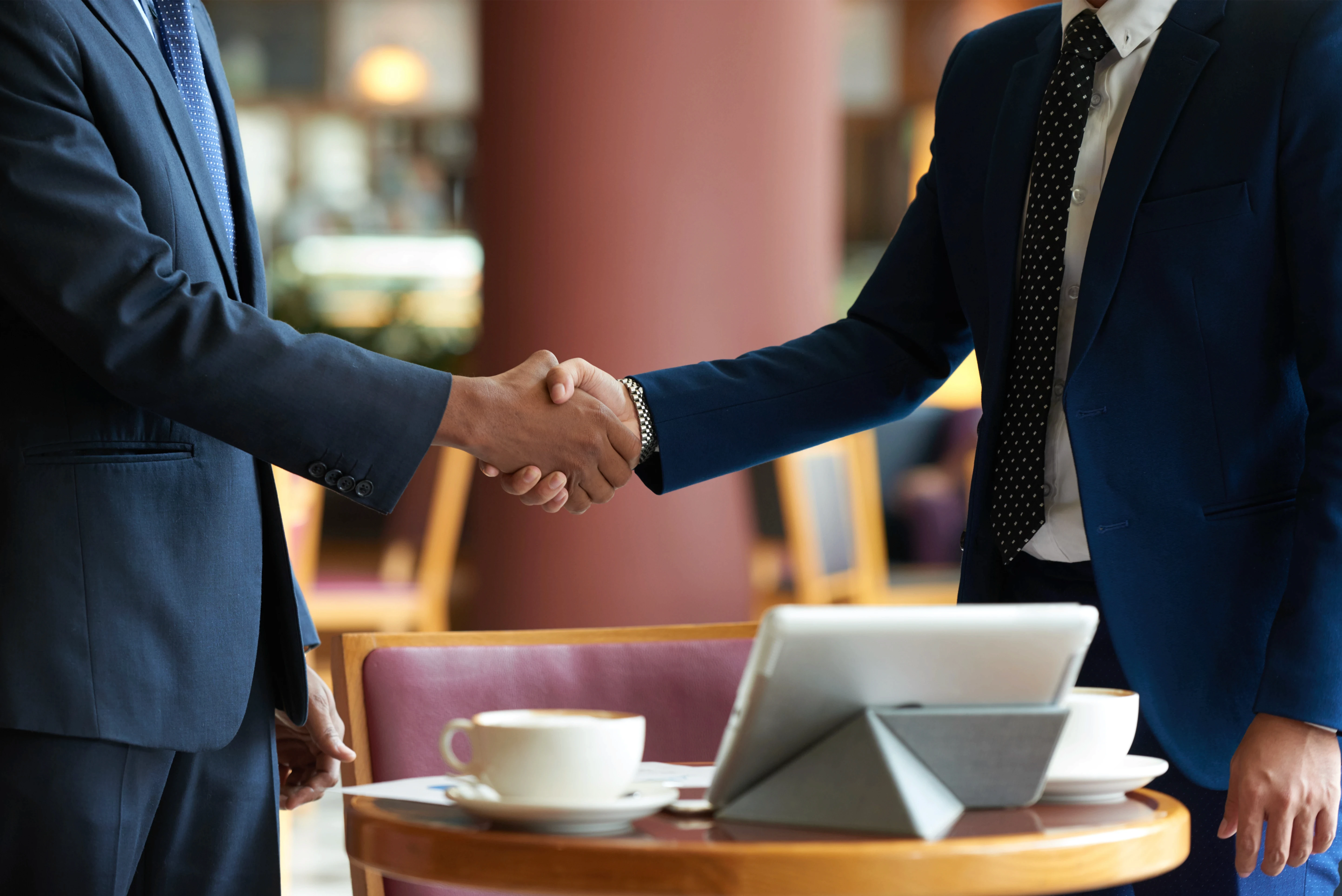 Two businesspeople in suits shake hands across a table in a cafe, with a tablet and coffee cup nearby, conveying a sense of agreement and professionalism.