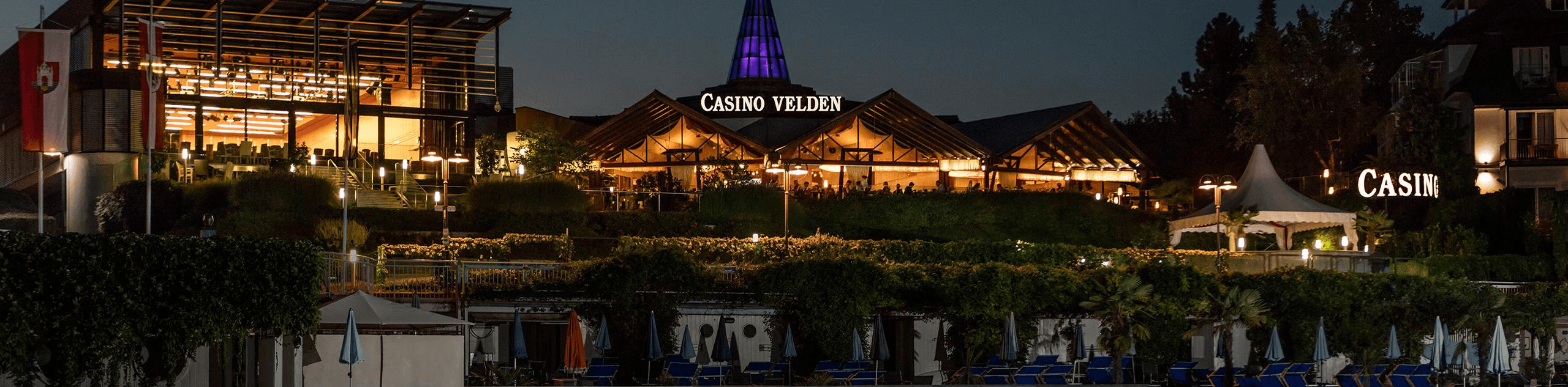 MW Interior  - Casino Velden am Wörthersee bei Nacht mit violetter LED-Beleuchtung und belebter Außenterrasse" Beschreibung: Das Casino Velden am Wörthersee – ein anspruchsvoller Referenzstandort, für den hochwertige Einrichtungslösungen im Hospitality-Bereich realisiert wurden.