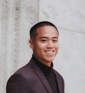 A person wearing a dark blazer and black turtleneck smiles against a backdrop of white, ornate architectural details, evoking professionalism and confidence.