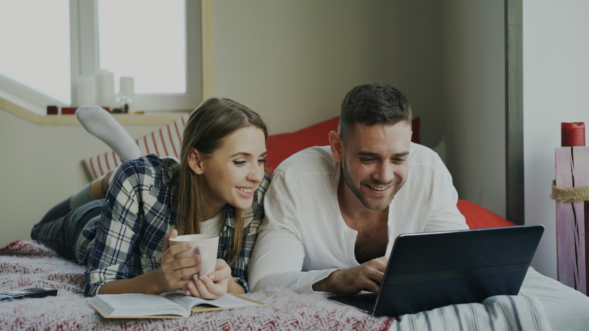 Couple lying on a bed with a mug of coffee, smiling at a laptop