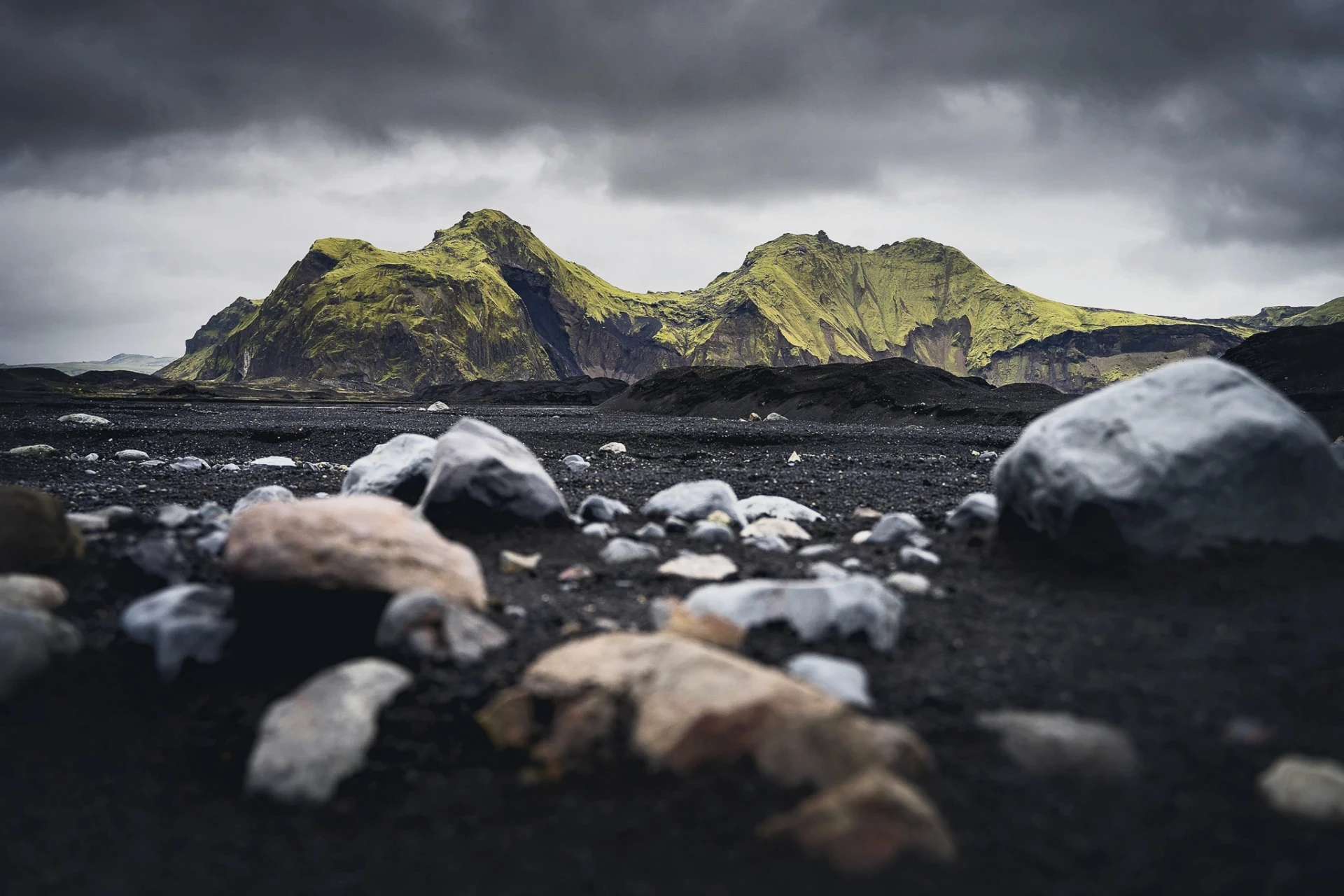 Moss-covered mountains under dark clouds with rocks on black sand.