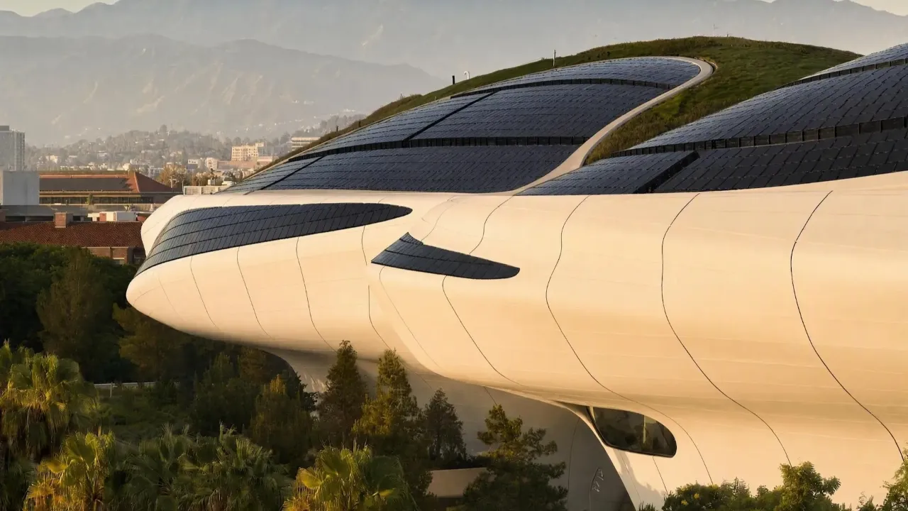 A modern, futuristic building with sleek, curved white structures and extensive solar panels on its roof surrounded by lush greenery and distant mountains under a clear sky.