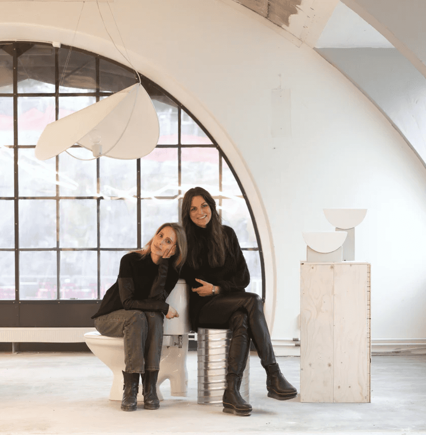 Two people dressed in black sit on a modern white chair in a bright, spacious room with a large, arched window, alongside minimalist white sculptures on a wooden pedestal.